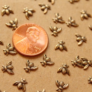 May include: A close-up of a group of small, gold-colored bee charms scattered on a brown surface. A US penny is in the center of the image for scale.