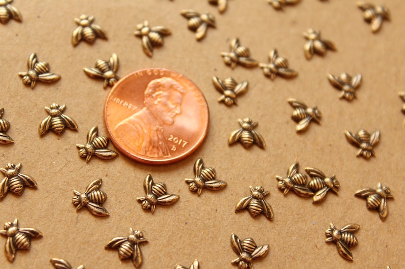 May include: A close-up of a group of small, gold-colored bee charms scattered on a brown surface. A US penny is in the center of the image for scale.