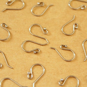 May include: Close-up of several silver-colored earring hooks scattered on a tan surface. Each hook has a curved design with a small ball at the end. The hooks are made of a shiny metal.