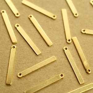 May include: A collection of gold-colored metal bar pendants with a loop at the top. The pendants are scattered on a brown surface.