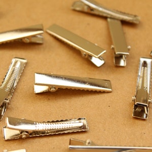 May include: A group of silver alligator hair clips scattered on a brown surface. The clips are all the same size and shape, with a serrated edge and a spring-loaded mechanism.