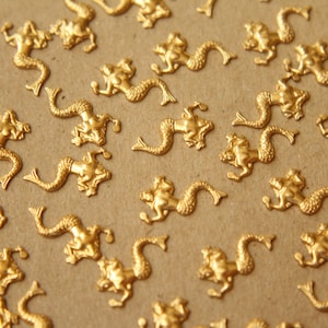 May include: A collection of small, gold-colored mermaid charms scattered on a neutral-toned surface. The charms are detailed with mermaid figures, featuring flowing tails and outstretched arms. These decorative charms are ideal for crafting and jewelry making.