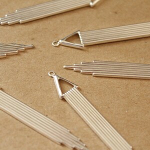 May include: Five silver metal pendant charms with a geometric design. Each charm features a series of thin, parallel lines that create a linear pattern. The charms have a triangular shape at the top and a pointed end at the bottom.