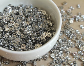 May include: A white ceramic bowl overflowing with shiny, silver-colored earring backs. The earring backs are scattered around the bowl on a light brown surface. The earring backs are small, with a butterfly-shaped design.