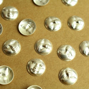 May include: A collection of silver-toned metal buttons with a raised design of a face. The buttons are arranged in rows on a brown surface.