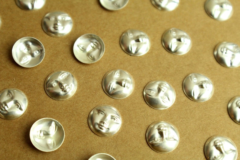 May include: A collection of silver-toned metal buttons with a raised design of a face. The buttons are arranged in rows on a brown surface.