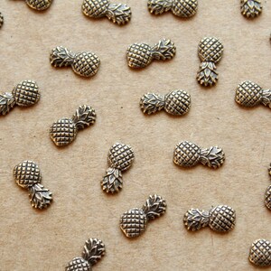 May include: A collection of gold-toned pineapple charms, scattered on a brown surface. The charms are small and detailed, with a realistic pineapple design.