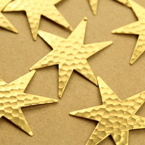 May include: A group of eight gold-toned metal star-shaped charms with a hammered texture. The stars are scattered on a brown surface.