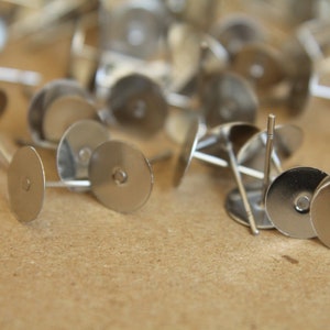 May include: A close-up of a group of silver earring backs. The backs are round and have a small hole in the center. They are scattered on a brown surface.