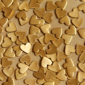 May include: A close-up image of a large quantity of gold heart-shaped metal charms. The charms are scattered on a brown surface.