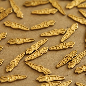 May include: A close-up of a group of gold-coloured carrot-shaped charms. The charms are scattered on a brown surface.