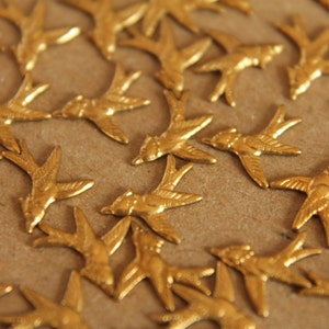 May include: A collection of gold-colored metal swallow bird charms. The charms are arranged in a random pattern on a brown surface.