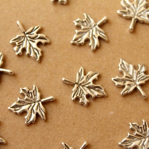 May include: A collection of silver maple leaf charms, scattered on a brown surface. The leaves are detailed and have a slightly textured finish.