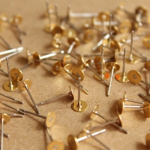 May include: A collection of gold-colored earring posts with silver-colored stems and flat, round backs. The posts are scattered on a light brown surface, with some in focus and others blurred.