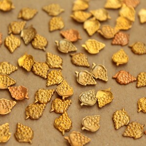May include: A collection of gold-colored metal leaf-shaped embellishments. The leaves have a textured surface and are scattered on a brown background.