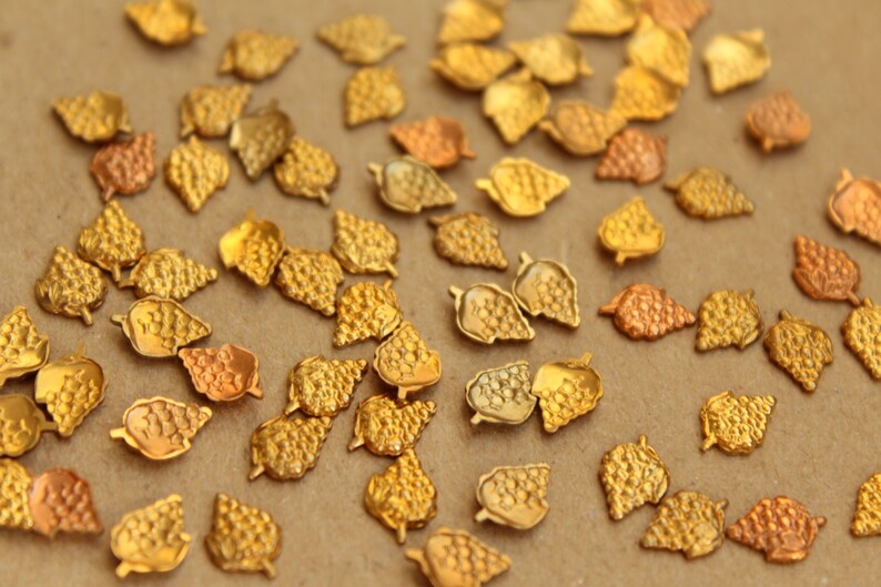 May include: A collection of gold-colored metal leaf-shaped embellishments. The leaves have a textured surface and are scattered on a brown background.