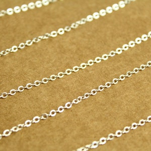 May include: Silver chain with a delicate, linked design. The chain is laid out in a diagonal pattern on a brown surface.