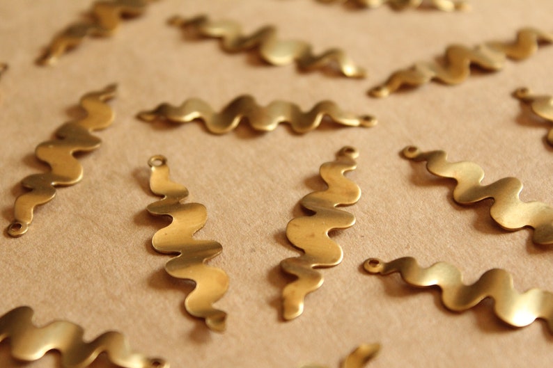 K&ouml;nnte beinhalten: Eine Sammlung von wellenf&ouml;rmigen Metallformen in Gold, jede mit einem leicht anderen Design. Die Formen sind auf einer braunen Oberfl&auml;che angeordnet.