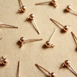 May include: A close-up of a group of 14 gold-toned earring posts with small ball-shaped tops. The posts are scattered on a brown surface.