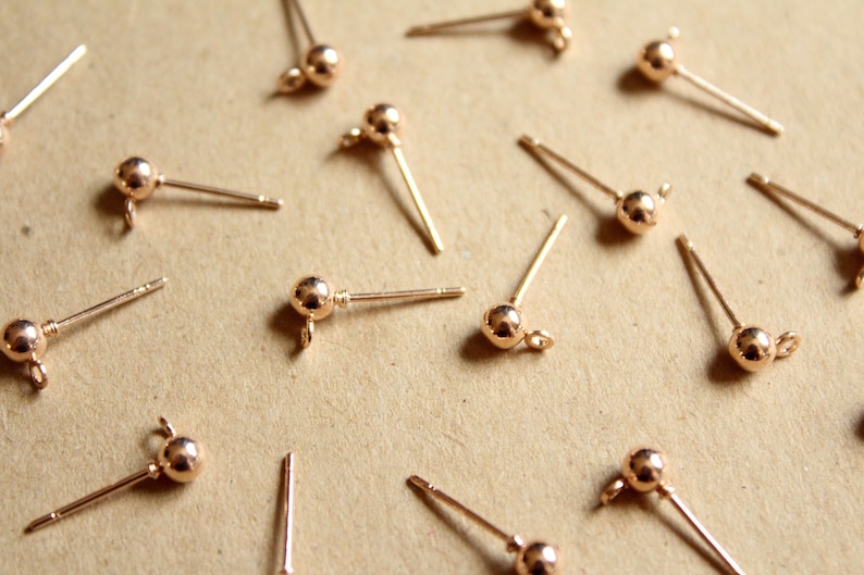May include: A close-up of a group of 14 gold-toned earring posts with small ball-shaped tops. The posts are scattered on a brown surface.