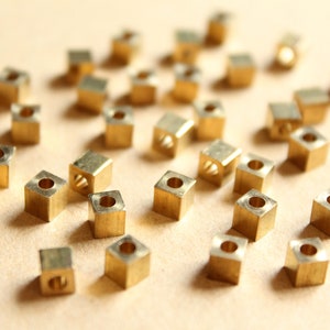 May include: A collection of small, gold-colored metal cubes with a hole in the center. The cubes are scattered on a light brown surface.