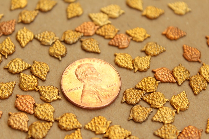 May include: A close-up of a group of gold-colored metal grape clusters. The clusters are scattered on a brown surface, with a US penny in the center for scale.