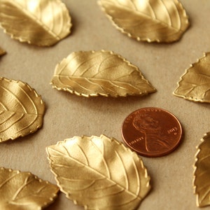 May include: A collection of ten gold-colored metal leaves with a textured surface. The leaves are scattered on a brown surface with a copper-colored penny in the center.