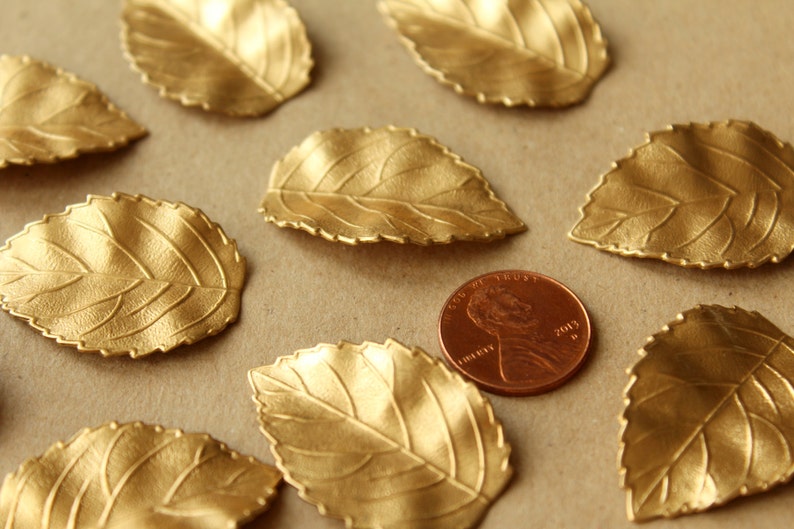 May include: A collection of ten gold-colored metal leaves with a textured surface. The leaves are scattered on a brown surface with a copper-colored penny in the center.