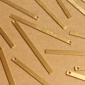 May include: Close-up of several small, rectangular, gold-colored metal bars with a small hole at one end. The bars are scattered on a light brown surface. These are likely jewelry making supplies.