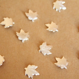 May include: A collection of silver maple leaf charms, scattered on a brown surface.