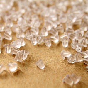 May include: A close-up of a pile of clear plastic earring backs. The backs are small and round, with a small hole in the center. They are scattered on a brown surface.