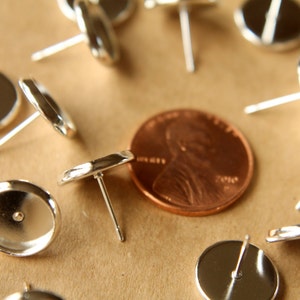May include: A close-up of a group of silver earring posts with round backs. The earring posts are arranged in a circle around a penny.