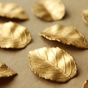 May include: A collection of gold-colored leaf-shaped metal embellishments. The leaves have a textured surface and are arranged on a brown background.
