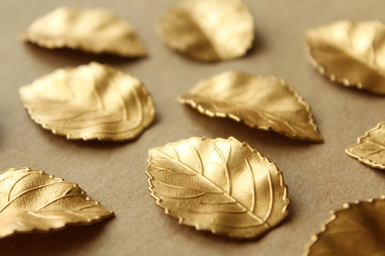 May include: A collection of gold-colored leaf-shaped metal embellishments. The leaves have a textured surface and are arranged on a brown background.