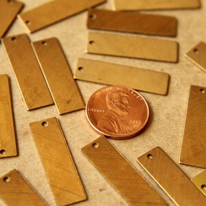 May include: A collection of rectangular brass tags with a small hole at one end, scattered on a neutral surface. A copper penny is included for size reference. The tags have a brushed metal finish.