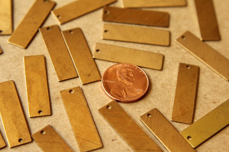 May include: A collection of rectangular brass tags with a small hole at one end, scattered on a neutral surface. A copper penny is included for size reference. The tags have a brushed metal finish.