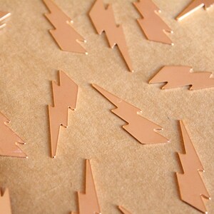 May include: A collection of small, copper-colored lightning bolt charms scattered on a brown surface. The charms are flat and have a shiny finish, ideal for jewelry making or craft projects. The bolts are all facing different directions.