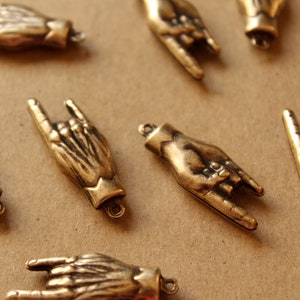 Puede incluir: Una colección de diez abalorios de metal dorado en forma de manos haciendo el gesto de rock and roll. Los abalorios tienen un acabado ligeramente envejecido y son perfectos para añadir un toque único a las joyas o las manualidades.