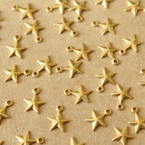 May include: A collection of gold-toned metal star charms with a loop on the top. The charms are scattered on a brown surface.