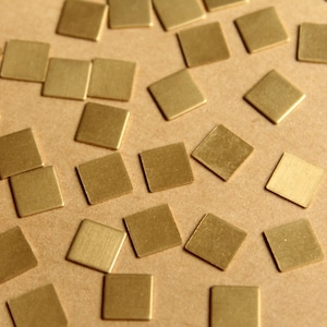 Könnte beinhalten: Eine Ansammlung kleiner, quadratischer, messingfarbener Metallfliesen, die auf einer hellbraunen Oberfläche verstreut sind. Die Fliesen haben eine matte Oberfläche und sind gleichmäßig verteilt, wodurch ein strukturiertes Aussehen entsteht. Diese dekorativen Fliesen könnten für verschiedene Bastelprojekte verwendet werden.