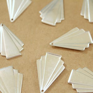 May include: A collection of silver-colored metal charms in the shape of a fan. The charms are arranged in a random pattern on a brown surface.