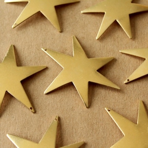 Puede incluir: Primer plano de varios adornos dorados en forma de estrella. Cada estrella tiene seis puntas y un pequeño agujero en la parte superior. Las estrellas están dispersas sobre una superficie marrón claro, creando un patrón.