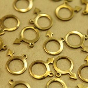 May include: A group of gold-colored metal charms in the shape of a circle with an arrow attached. The charms are scattered on a brown surface.