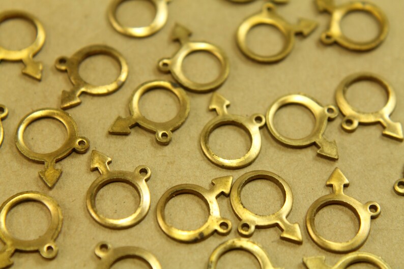 May include: A group of gold-colored metal charms in the shape of a circle with an arrow attached. The charms are scattered on a brown surface.