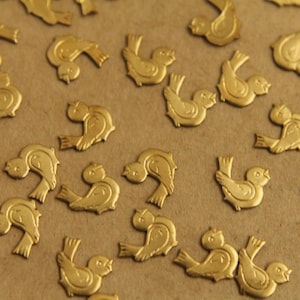 Puede incluir: Una dispersión de abalorios de pájaros de metal dorado sobre una superficie marrón. Los pájaros están todos mirando en la misma dirección y tienen un diseño simple y estilizado.