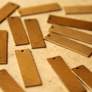 May include: A collection of small, rectangular brass tags scattered across a surface. Each tag has a single hole at one end, and a brushed finish. The tags are a warm, golden color, and are likely used for jewelry making or crafting.