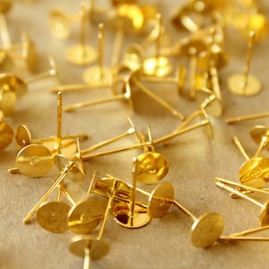 May include: A close-up of a group of gold earring posts with flat backs. The posts are scattered on a brown surface.