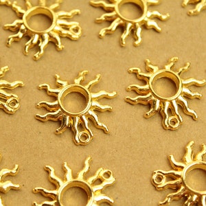 May include: Gold-colored sun-shaped charms with a ring in the center and radiating rays. These metal charms have a small loop at the bottom for attaching to jewelry or other crafts. The charms are arranged on a tan surface.