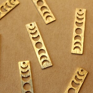 May include: Eight gold-colored metal charms with a moon phase design. Each charm is rectangular and has a hole at the top for hanging.