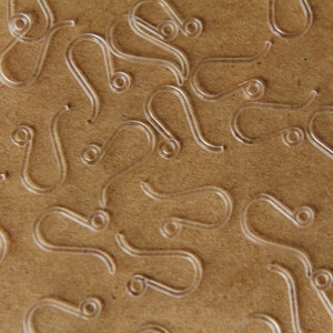 May include: A close-up image of a pile of clear plastic earring hooks. The hooks are scattered on a brown surface.